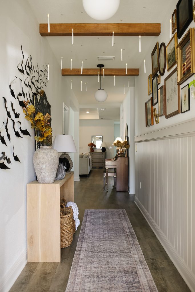 cozy fall entryway- looking through a front door at a long hallway with wooden beams on the ceiling, shiplap on half the right side wall with a gallery wall of photos above. There's a console table on the left wall surrounded by decorative bats going up the wall. floating candles are hanging from the ceiling